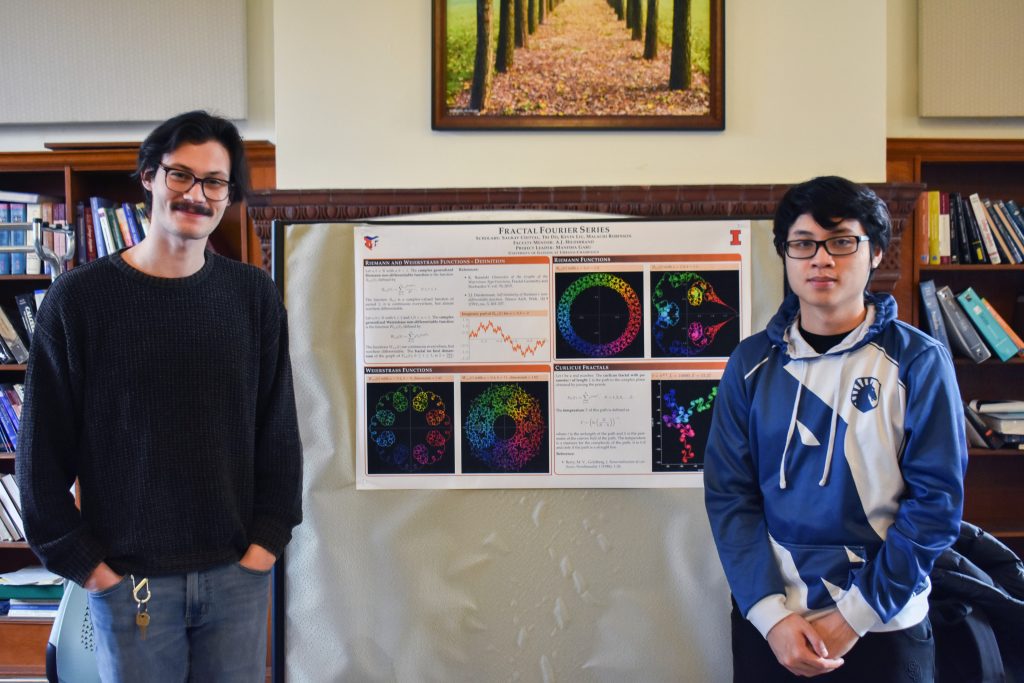 Two students stand in front of a research poster in a library. The poster is titled "Fractals in Fourier Series>"